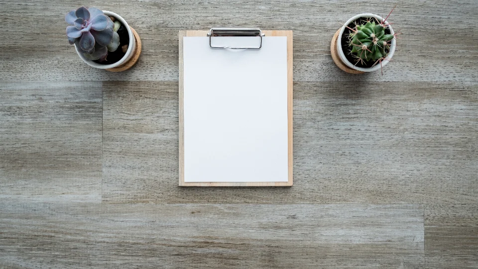 A blank sheet of paper on a clipboard between two small potted plants on a wooden desk