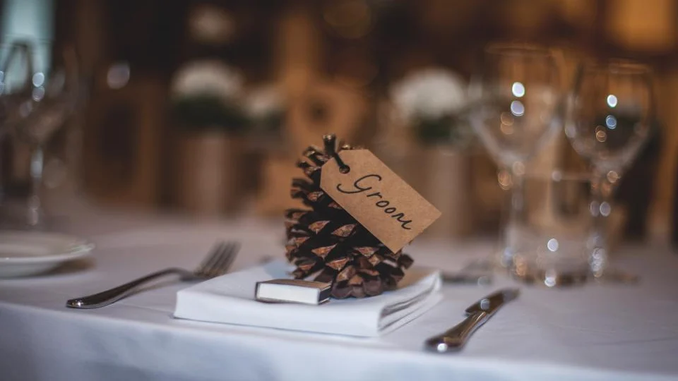 A handwritten place card tied to a pine cone, on a napkin at a table laid for a meal