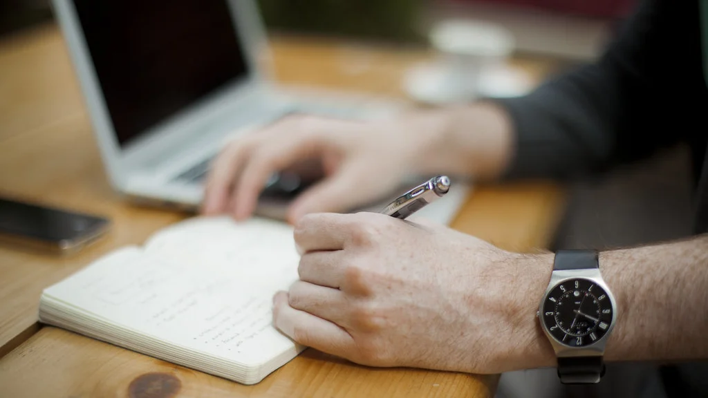 A person writing in a notebook beside an open laptop