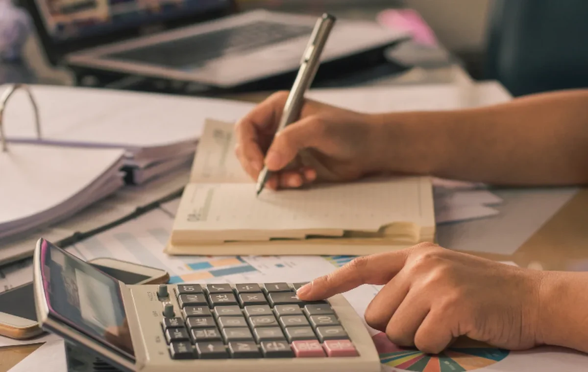 A hand writing figures on paper beside a calculator